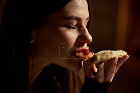 Una hermosa chica con los ojos cerrados disfruta comiendo pizza. Los labios están pintados con lápiz labial rojo.
