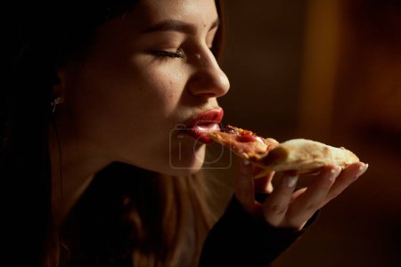 Una hermosa chica con los ojos cerrados disfruta comiendo pizza. Los labios están pintados con lápiz labial rojo.