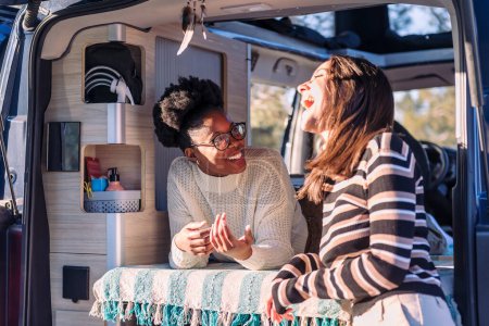 two happy women laughing in a conversation and enjoying van life, concept of weekend getaway with best friend and relaxation in camper van