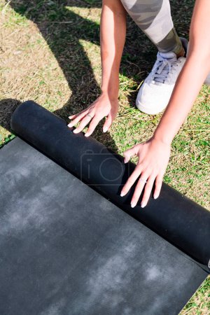 Téléchargez les photos : Mains d'une jeune femme méconnaissable tapis roulant pour une séance de yoga sur l'herbe dans le parc, concept de mode de vie sain et actif - en image libre de droit