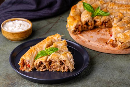 Piece of Rolled Burek - savoury round pie fith meat, onion, tomato, bell pepper feeling, decorated with sesam seedes, yogurt. Dark green table surface. 