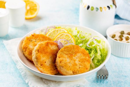 Fish cakes or burger, or cutlets. Made from ground perch and tuna with herbs, breaded and fried, served with iceberg salad, lemon slices. 