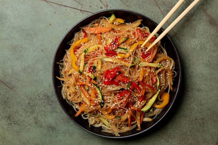 Téléchargez les photos : Top view of a plate with Japchae , Korean glass noodle made with sweet potato starch,  stir fried with vegetables.  Zucchini, bell pepper, carrot, sesame seed. Green table surface. - en image libre de droit