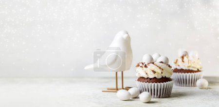 Photo for Beaitiful festive background. Chocolate cupcakes with cream cheese nest, egg shaped sweet candies and grated chocolate.  Bird statue. Holiday homemade dessert. Copy space for your text, banner size. - Royalty Free Image