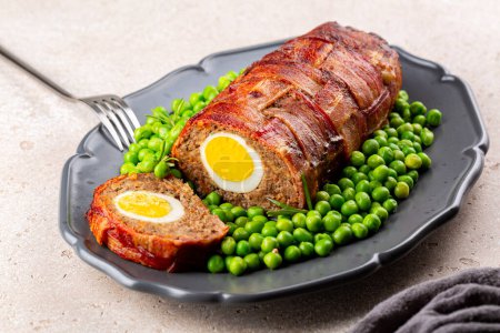 Photo for Slice of baked meatloaf made of ground meat, onion, carrot, stuffed with hard-boiled eggs, wrapped with bacon net, served with green peas. German, Scandinavian and Belgian dish. Festive and Easter food. - Royalty Free Image