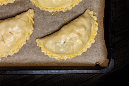Photo for Cooking meat turnovers or pies, or empanadas, or cornish pasty. Unbaked shortcrust pastry,  egg washed, on a baking tray. - Royalty Free Image