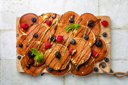 Photo for Pancakes board with chocolate and pistachio topping and fresh berries, blueberry, raspberry, boysenberry on a wooden board and white table surface. Directly above, top view. - Royalty Free Image
