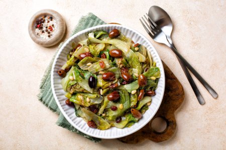 Photo for Italian salad made with sauteed broad-leaved endive or escarole with taggiasca olives, pine nuts, raisins. Vegan dish. Top view. - Royalty Free Image
