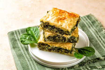 Stack of Spanakopita piece of pie, homemade Greek savory spinach pastry. It contains cheese feta, chopped spinach, green, egg, layered in phyllo or filo dought. Light beige background with green napkin.