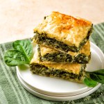 Stack of Spanakopita piece of pie, homemade Greek savory spinach pastry. It contains cheese feta, chopped spinach, green, egg, layered in phyllo or filo dought. Light beige background with green napkin.