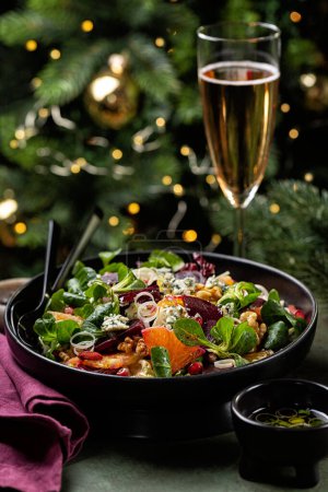 Photo for Christmas or New Year table. Winter salad with beetroot, oranges, walnuts, pomegranate, dried cranberries, valerian lettuce, blue cheese. Pink wine in glass. Honey and olive oil dressing. Christmas tree on background. - Royalty Free Image