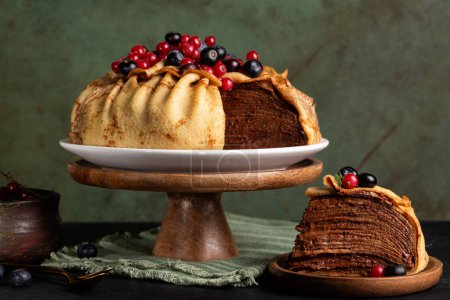 Homemade layered crepe cake, made of type of thin pancake and chocolate cream, decorated with blueberry and red currant, served on a dark table, green background.