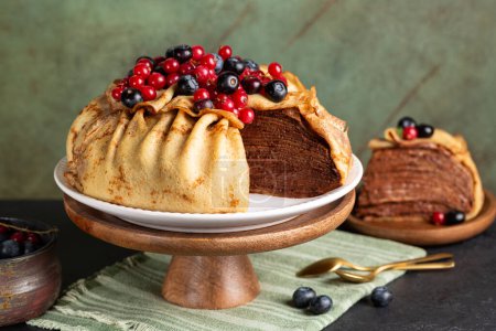 Homemade crepe cake, made of type of thin pancake and chocolate cream, decorated with blueberry and red currant, served on a dark table, green background.