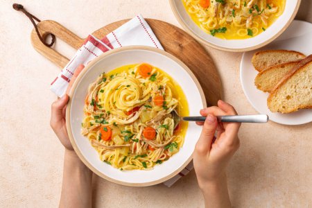 Photo for Eating chicken noodle soup in a white plate, on a wooden board. Girls hands holding spoon. Directly above on a beige table. - Royalty Free Image
