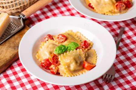Photo for Plate with creamy Italian Ravioli with tomatoes and cheese. Table with traditional red and white chess tablecloth. - Royalty Free Image
