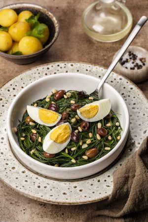 Frühlingssalat. Agretti mit Taggiasca-Oliven, Pinoli oder Pinienkernen und gekochten Eiern anbraten. Vertikales Bild.