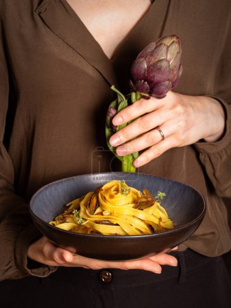 Frau hält einen Teller mit Artischocken-Nudeln, Fettucine, gewürzt mit Safran und Thymian. Frische essbare Artischockenblume in der Hand. Italienisches Ernährungskonzept.