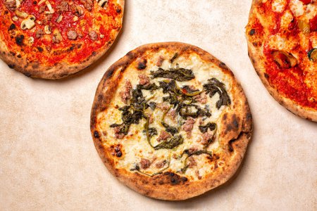 Top view of three different Italian pizza. White Pizza with cheese, sausages and Rapini or broccoli rabe, red pizza with salsiccia and mushrooms, and classic peperoni. Beige table surface.
