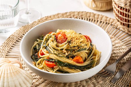 Spaghetti with agretti, or salsola soda, tomatoes, bread crumbs, pinoli pine nuts and anchovy in a white plate. Italian pasta, white dinner table.
