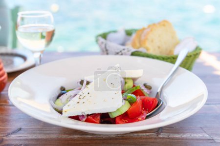 Téléchargez les photos : Traditional Greek Salad served in tavern, traditional greece food with Aegean sea as background. Tomatoes, cucumber, onions, olives, peppers, cappers and olive oil - en image libre de droit