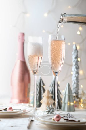 Weihnachten Tischdekorationen in weiß und rosa Farben mit trendigen Papier Weihnachtsbäume auf dem Hintergrund. Tisch bereit für festliches Abendessen und Party mit rosa Prosecco im Glas