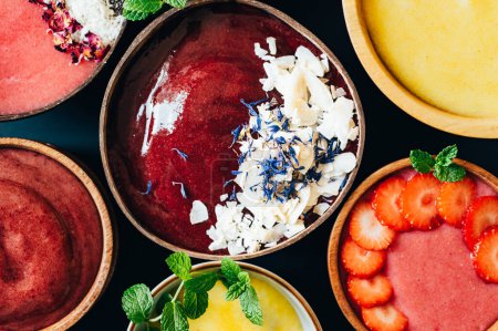 Photo for Variety of different smoothies: Banana and Strawberries, Cherry and Apple, Pineapple and Mango with pieces of Strawberries, Coconut Shreds, Chia Seeds and rose petals on top in the Coconut Shell Bowl - Royalty Free Image