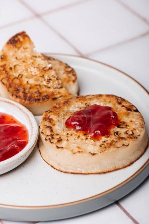 Hot Toasted Crumpets mit Butterstück auf einem Teller mit Erdbeermarmelade. Perfekt leckeres Frühstück beliebt in England, Großbritannien