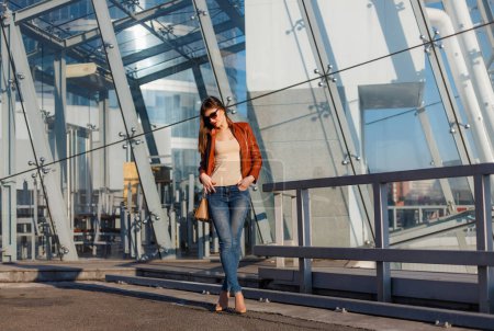 Photo for Portrait of beautiful brunette young woman in nice red brown jacket, denim jeans and sunglasses. Genuine leather bag, high heels shoes. Spring autumn fashion trendy photo of lady on urban background. - Royalty Free Image