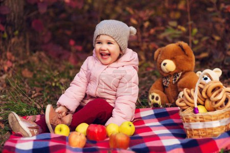 Téléchargez les photos : Portrait of beautiful happy child sitting on plaid with autumn colorful leaves background. Picnic basket with apples and bagels, toy bears. Funny girl outdoors in fall park. - en image libre de droit