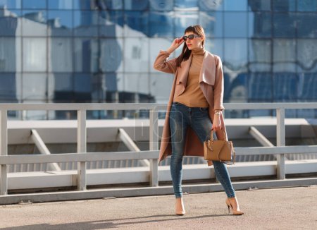 Photo for Portrait of beautiful brunette young woman in nice brown beige coat, denim jeans and sunglasses. Genuine leather bag, high heels shoes. Spring autumn fashion trendy photo of lady on urban background. - Royalty Free Image