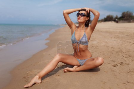 Photo for Portrait of sexy beautiful tanned woman posing in fashion striped retro style swimwear bikini and sunglasses at the sea coast. Exotic country travel and rest concept. Slim figure and sexy body. - Royalty Free Image