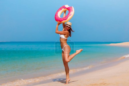 Téléchargez les photos : Vacances touristiques femme sautant et jouissant avec beignet rose drôle en bikini et chapeau. Modèle de plage riant à Dubaï. Voyage femme dans la célèbre destination EAU. Belle plage de sable eau claire à Jumeirah Dubaï. - en image libre de droit