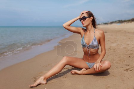 Photo for Portrait of sexy beautiful tanned woman posing in fashion striped retro style swimwear bikini and sunglasses at the sea coast. Exotic country travel and rest concept. Slim figure and sexy body. - Royalty Free Image