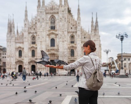 Foto de La turista de viaje alimenta a las palomas cerca del Duomo di Milano, la iglesia catedral de Milán en Italia. Chica disfrutando en la plaza en el centro de la ciudad, famoso destino para el viajero - Imagen libre de derechos
