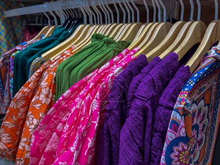 Auf einem Kleiderständer sind eine Vielzahl dynamischer Damenoberteile fein säuberlich ausgestellt. Die Oberteile hängen senkrecht und präsentieren ihre unterschiedlichen Farben und Muster. Der Ständer ist gut organisiert, wobei jedes Hemd gleichmäßig vom nächsten Hemd entfernt ist..