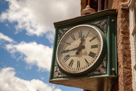 An ornate vintage clock mounted on a brick wall, displaying time under a clear blue sky with wispy clouds