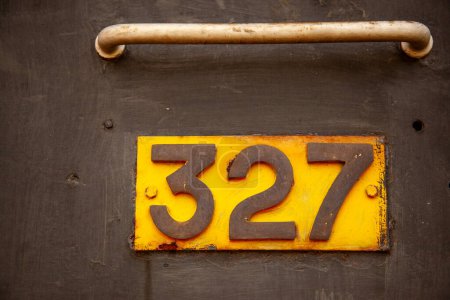 Primer plano de una placa metálica oxidada de color amarillo y naranja envejecida con el número 327 en una puerta con textura