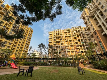 Wide angle view of apartment buildings surrounded by trees having a playground lawn in the center with some chairs lying around. With blue sky in the background.