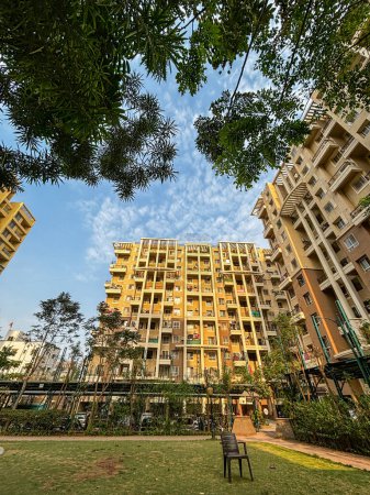 Wide angle view of apartment buildings surrounded by trees having a playground lawn in the center with some chairs lying around. With blue sky in the background.
