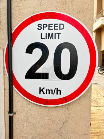In Nahaufnahme eines Schildes mit Geschwindigkeitsbeschränkungen, das an einer cremefarbenen Wand angebracht ist, zeigt die Tafel eine Geschwindigkeitsbegrenzung von 20 km / h an. Eine schwarze Stange läuft senkrecht über die Ecke des Boards.