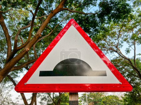 A red and white sign with a black triangle in the middle. The sign is pointing to a road with a speed breaker/dip in it