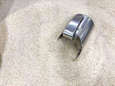 Une cuillère en argent est posée sur un tas de riz blanc. La cuillère est pliée et a un trou dedans. Le riz est étalé sur le sol, et la cuillère est le seul objet visible. La scène est simple