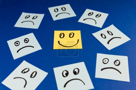 Klebezettel mit handgezeichnetem Smiley-Gesicht und traurigen Gesichtern auf blauem Hintergrund. Positives Denkkonzept. Positiv überwindet Negatives