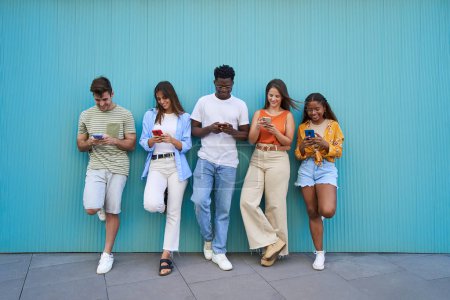 Photo for Multicultural group of friends using cell phones - Students leaning against a blue wall and typing on smartphones. - Royalty Free Image