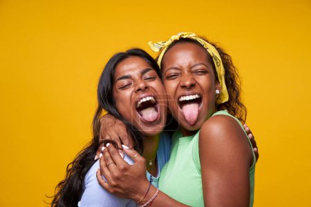 Foto de Two diversity pretty positive girls stick out your tongue hugging isolated on yellow color background. Young couple goofing around embracing and looking camera. Friendship and homosexual relationship. - Imagen libre de derechos