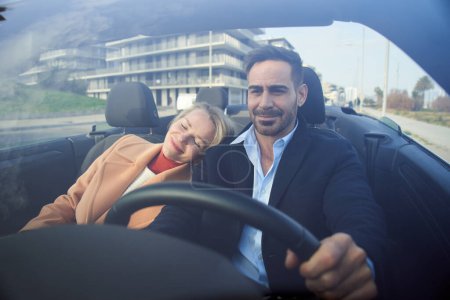Photo for Middle aged couple riding convertible car on urban road. Happy blonde woman leaning on attractive man enjoying vacation feeling of freedom. People driving carefree in landscape with city in background - Royalty Free Image