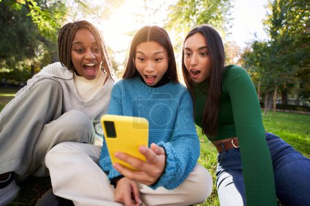 Foto de Tres mujeres jóvenes usando un teléfono celular en el parque. Retrato de chicas adolescentes sorprendidas mirando las redes sociales en el móvil. Gente mirando la pantalla del teléfono fascinada con la boca abierta. - Imagen libre de derechos