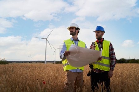 Foto de Dos ingenieros en sombreros duros trabajan sosteniendo el avión en el campo agrícola con turbinas eólicas. Técnicos profesionales en exteriores de central eólica. Concepto de energía limpia renovable, futuro sostenible. - Imagen libre de derechos