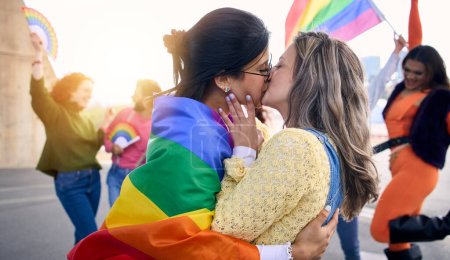Deux jeunes filles aimantes qui s'embrassent le jour du festival gay pride en plein air. Groupe d'amis célébrant fête lgbt sur fond avec drapeau arc-en-ciel et les fans. Génération z et types de sexualité.