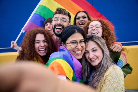 Téléchargez les photos : Selfie d'un groupe LGBT de jeunes célébrant la journée de la fierté gay tenant ensemble le drapeau arc-en-ciel. Communauté homosexuelle souriant et prenant autoportrait gai. Lesbiennes couple et amis génération z - en image libre de droit